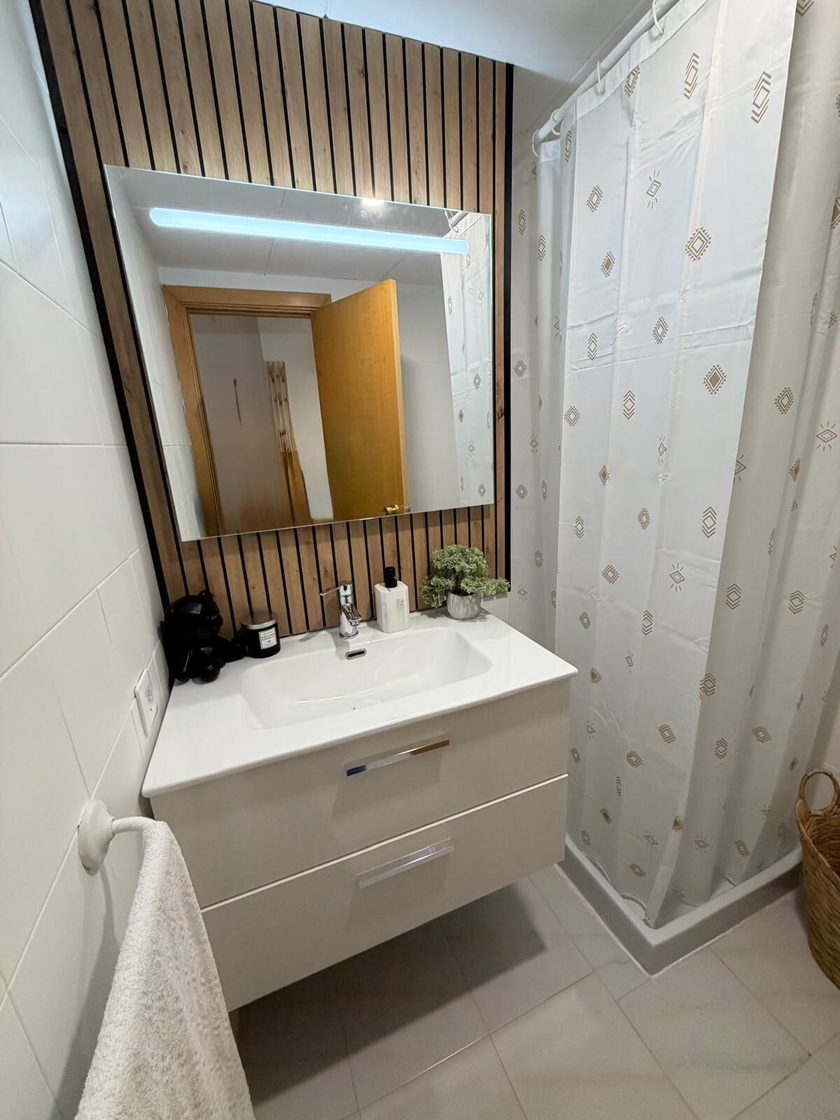 Een moderne badkamer met een wit wastafelmeubel, een grote spiegel, een douchegordijn met een geometrisch patroon en een kleine potplant op het wastafelmeubel.