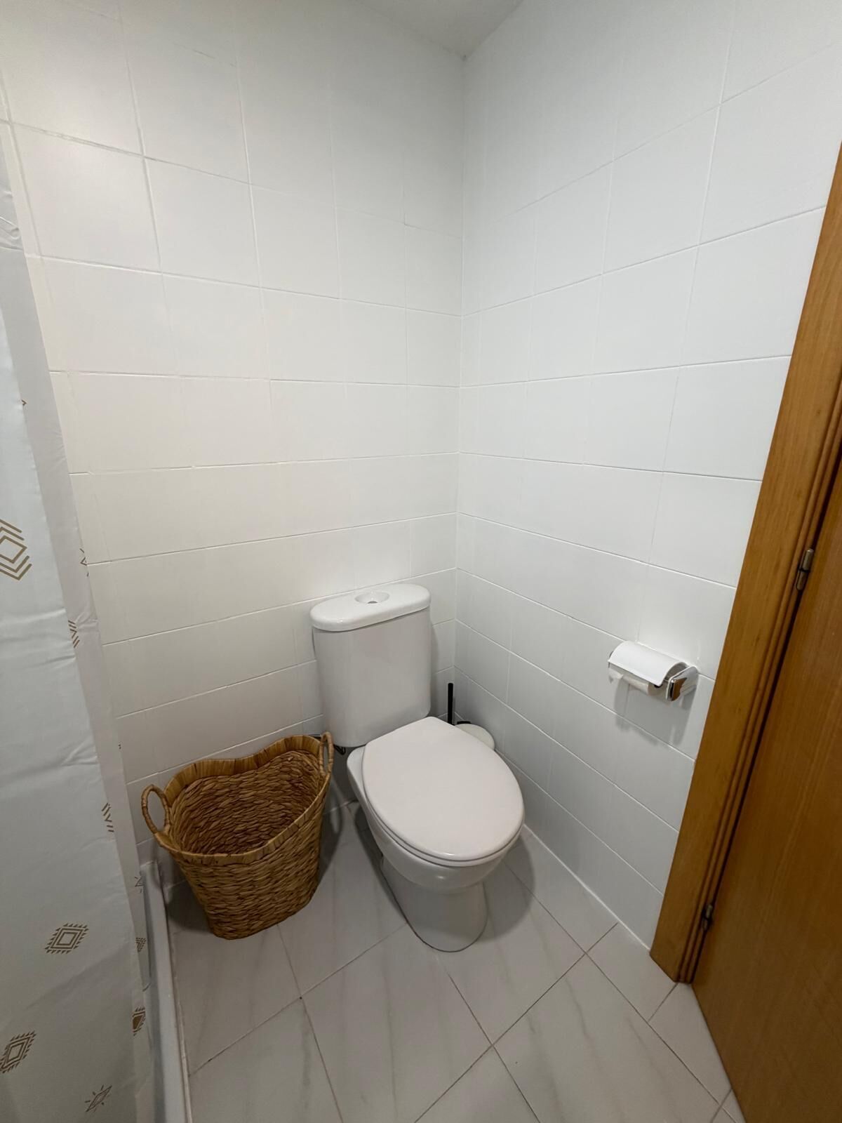 A small, clean bathroom with white tiles, a toilet, a toilet paper holder, and a wicker basket.