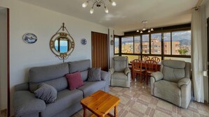 Ein Wohnzimmer mit einer Couch, Sesseln, einem Holztisch, einem Esstisch mit Stühlen, einer Deckenleuchte und einem großen Fenster mit Blick auf die Stadt.