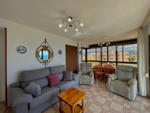 A living room with a sofa, armchairs, a wooden coffee table, a dining table with chairs, a ceiling light fixture, and a large window with a city view. - TERRASOL SAN GERARDO ON THE SEA (Torre del Mar)