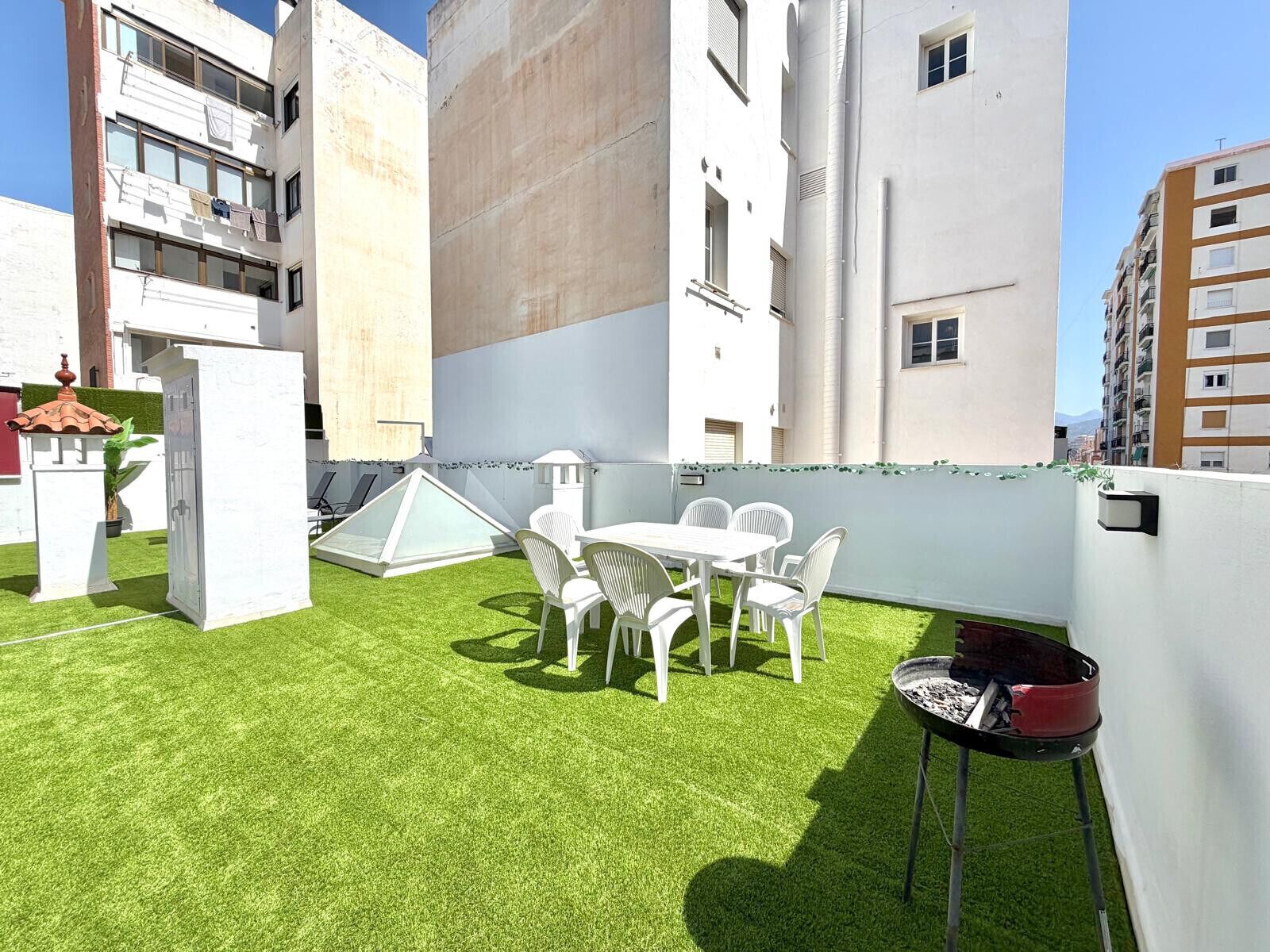 Une terrasse sur le toit avec une table et des chaises blanches, un barbecue et un petit espace jardin agrémenté de gazon synthétique.