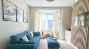 Ein Wohnzimmer mit einer Couch, einem kleinen Tisch, einem Fernseher und einem Fenster mit Vorhängen.