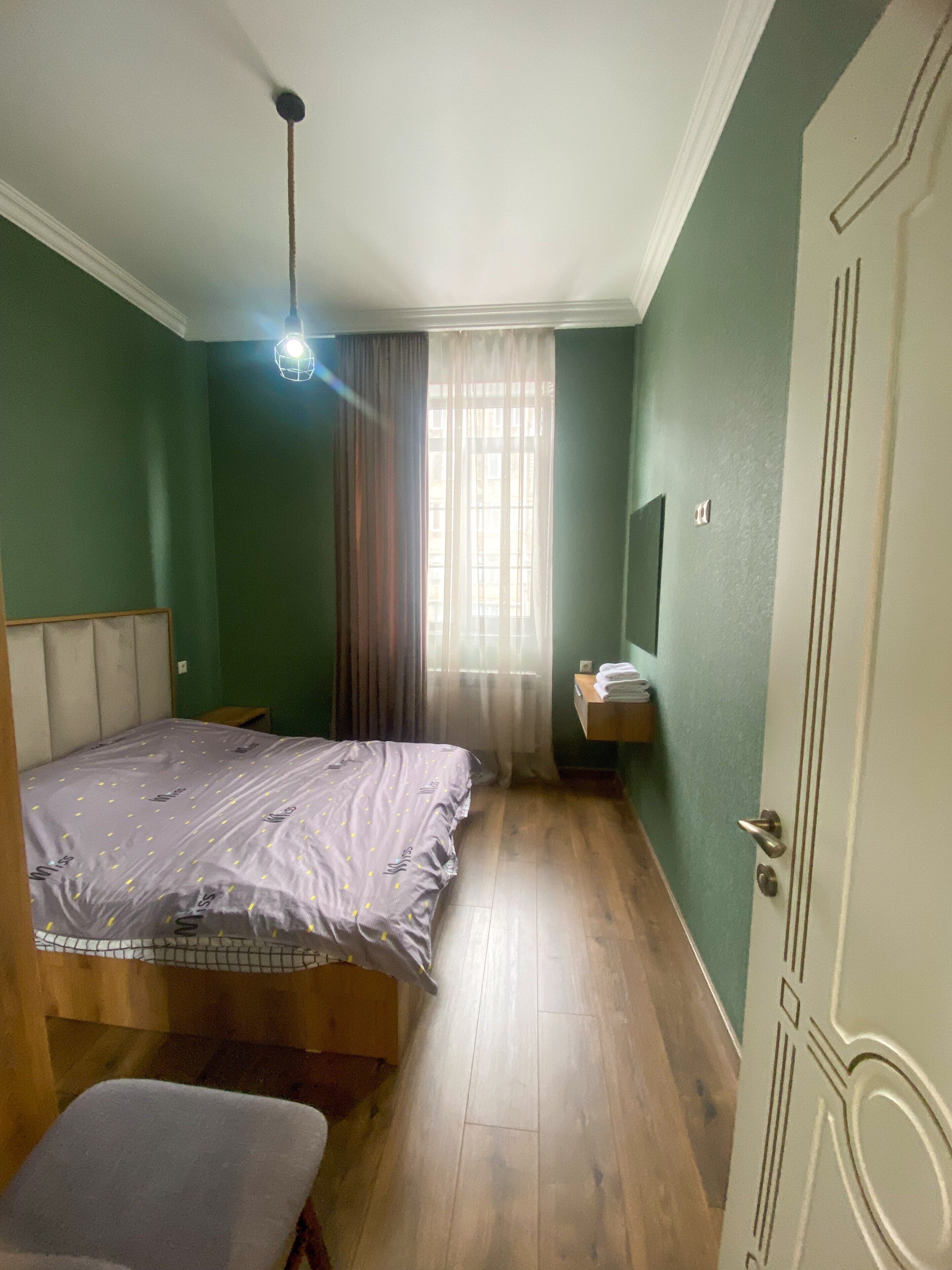 Ein Schlafzimmer mit einem Bett, einer Bank, einem Nachttisch, einem Fenster mit Vorhängen und einer Tür.