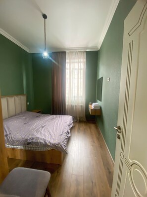 A bedroom with a bed, a bench, a bedside table, a window with curtains, and a door.