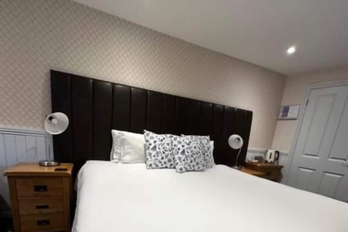 Una habitación de hotel con una cama grande, cabecera, mesita de noche, lámpara, cómoda y alfombra estampada.