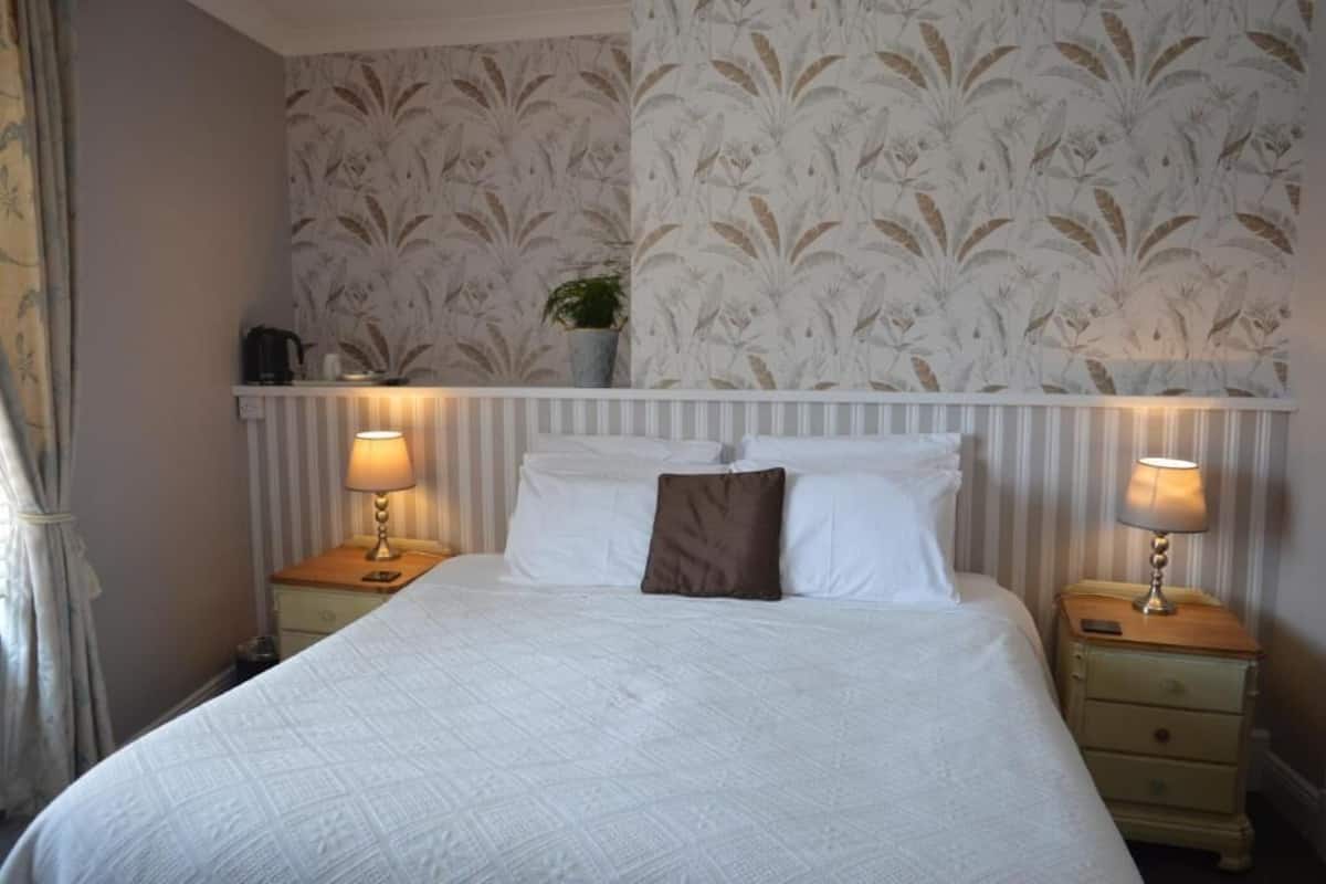 Una habitación de hotel con una cama grande, dos mesitas de noche con lámparas, un cabecero blanco y papel tapiz con motivos florales.