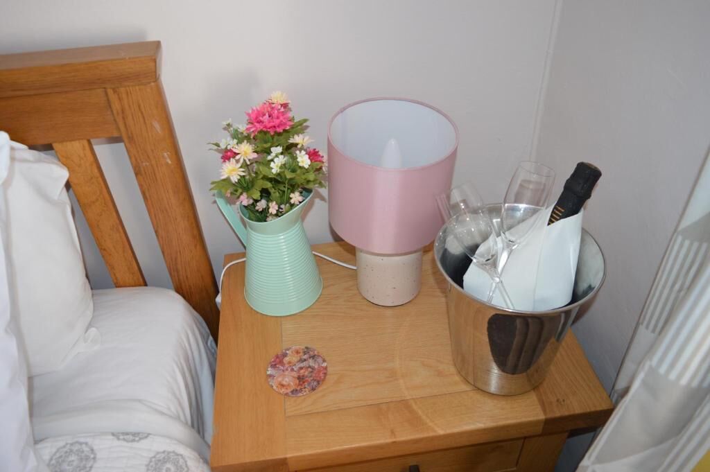 Una mesita de noche de madera con un florero turquesa lleno de flores, una lámpara rosa y un recipiente metálico con vasos y una botella.