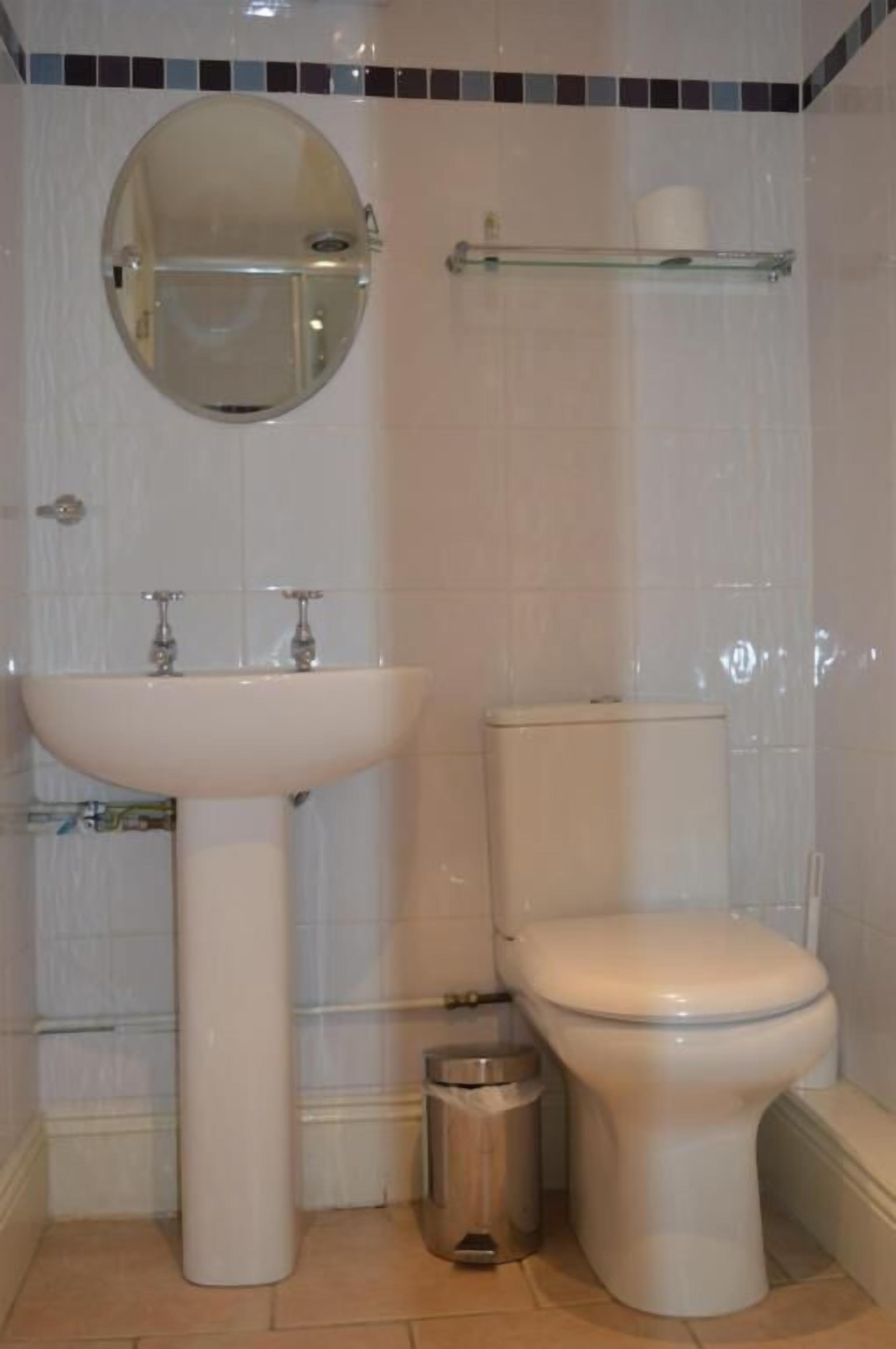 A bathroom with a pedestal sink, a toilet, a trash bin, a mirror, and a toilet paper holder.