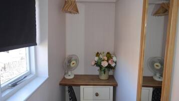 A small room with a window, a wooden shelf with a fan and flowers, a mirror, and a white dresser with drawers.