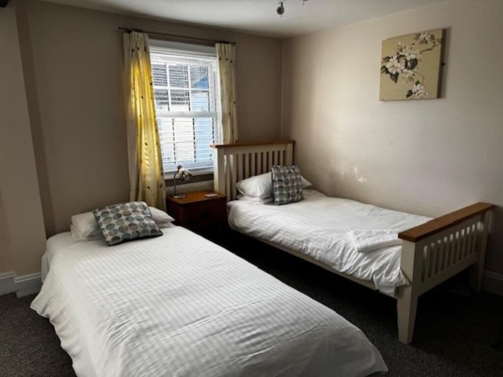 Un dormitorio con dos camas individuales, una ventana con cortinas, una mesita de noche y una obra de arte colgada en la pared.