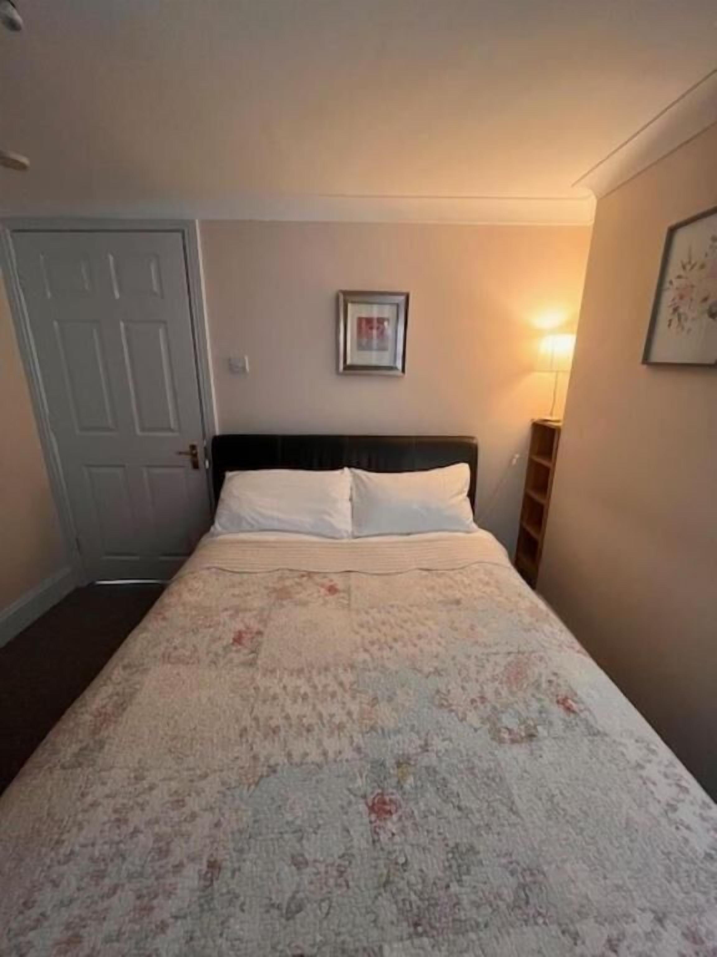 Un dormitorio con cama, una puerta, un cuadro enmarcado en la pared y una mesita de noche con lámpara.
