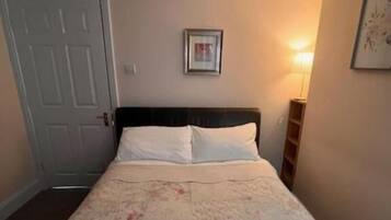 Un dormitorio con cama, una puerta, un cuadro enmarcado en la pared y una mesita de noche con lámpara.