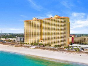Ein großes, mehrstöckiges Hotel mit gelber Fassade, direkt neben einem Strand mit klarem, blauem Wasser.