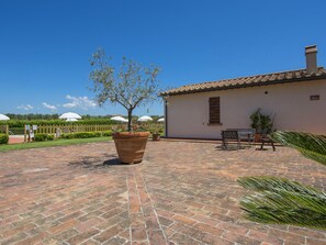 Une cour avec un sol en briques, un grand arbre planté dans un pot, un bâtiment avec un toit en tuiles et des sièges à l’extérieur.