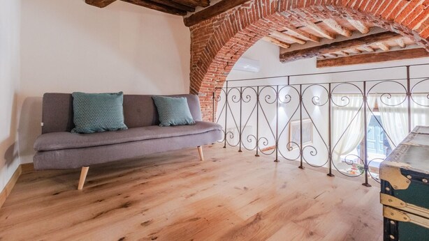 Habitación estilo loft con ladrillo visto, techo de madera, sofá gris con cojines estampados y barandilla de balcón de hierro forjado.