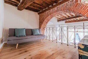 A loft-style room with exposed brick, a wooden ceiling, a grey sofa with patterned cushions, and a wrought iron balcony railing. - Antica Tipografia in Lucca (Lucca)
