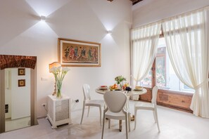 A dining area with a table set for four, a white chair, a decorative cabinet, a painting on the wall, and a window with curtains. - Antica Tipografia in Lucca (Lucca)