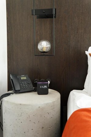 Une chambre d’hôtel avec une table d’appoint ronde et texturée, un téléphone noir, une horloge numérique affichant 11 h 29, et un objet décoratif encadré placé au-dessus de la table.