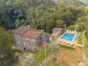 A stone building with a tiled roof, a swimming pool with lounge chairs, and a forested backdrop. - 7BR Tuscan Oasis with Pool_Borgo sulla Visona (Capannori)
