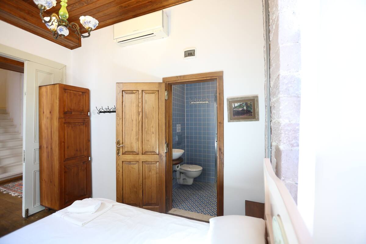 Un dormitorio con un armario de madera, una cama con ropa blanca y un baño con cabina de ducha azulejada en azul.
