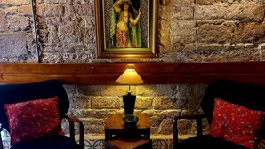 A room with a stone wall, a framed painting, two chairs with red pillows, a table with a lamp, and patterned floor tiles. - Kidalyo Hotel (Ayvalik)