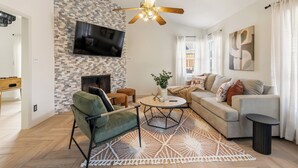 A modern living room with a fireplace, a flat-screen TV, a ceiling fan, and a large window with curtains. - Berrywood Stay / Pool + Fire Pit + Foosball (Garland)