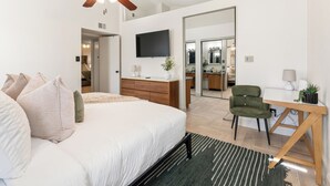 A modern bedroom with a bed, a desk, a chair, a television, and a bathroom visible through an open door. - Berrywood Stay / Pool + Fire Pit + Foosball (Garland)