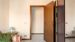 Una habitación con sofá, una mesita de noche con lámpara, un armario de madera y una puerta que da a otra estancia.