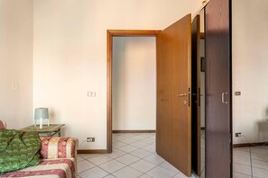 A room with a sofa, a small table with a lamp, a wooden wardrobe, and a door leading to another room. - Three Bedroom Apartment (Montecatini Terme)