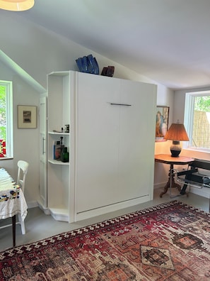 A room with a white wardrobe, a table with a lamp, a patterned rug, and a chair.