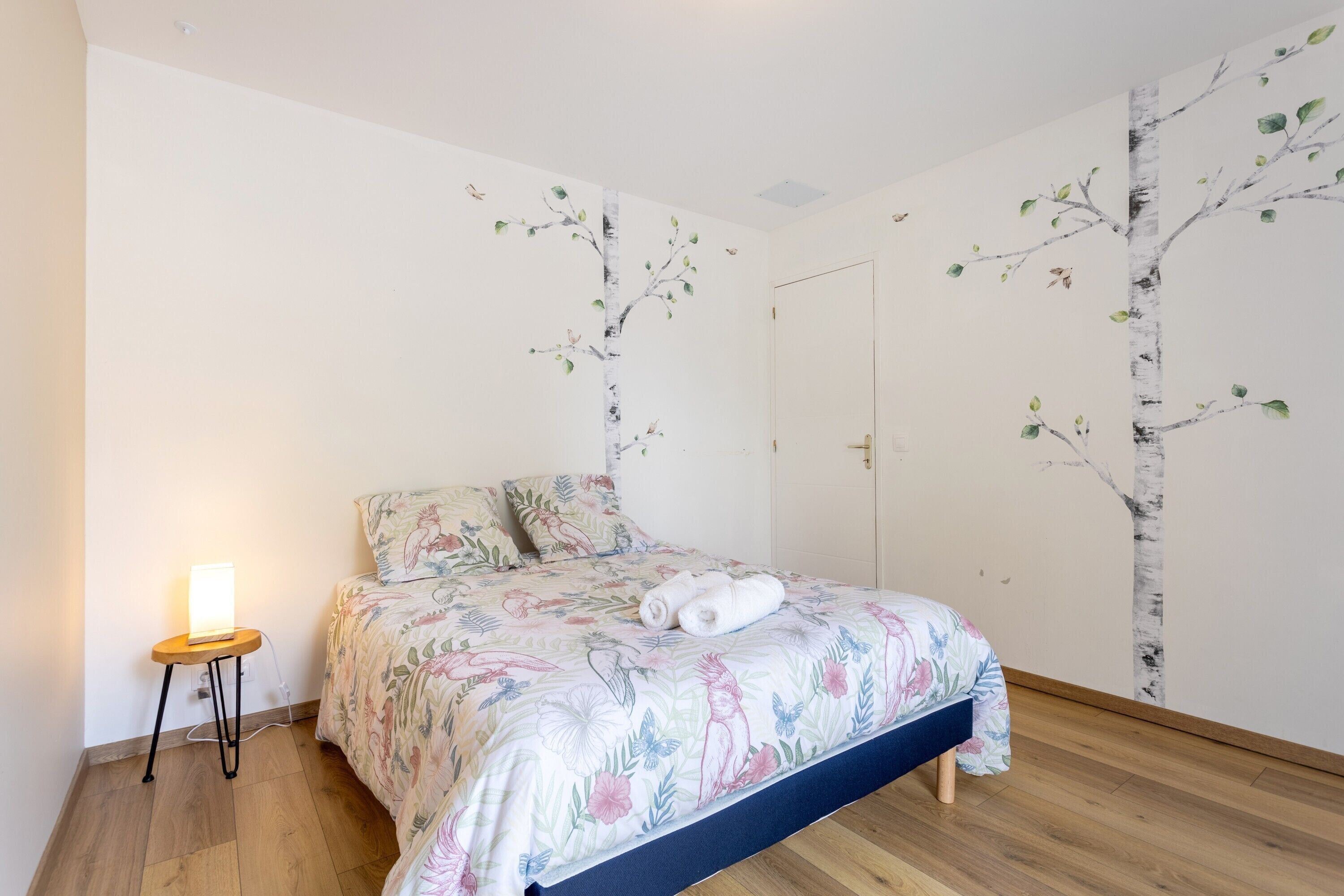 Een slaapkamer met een bed, een nachtkastje met een lamp, en een muur met een muurschildering van een boom.