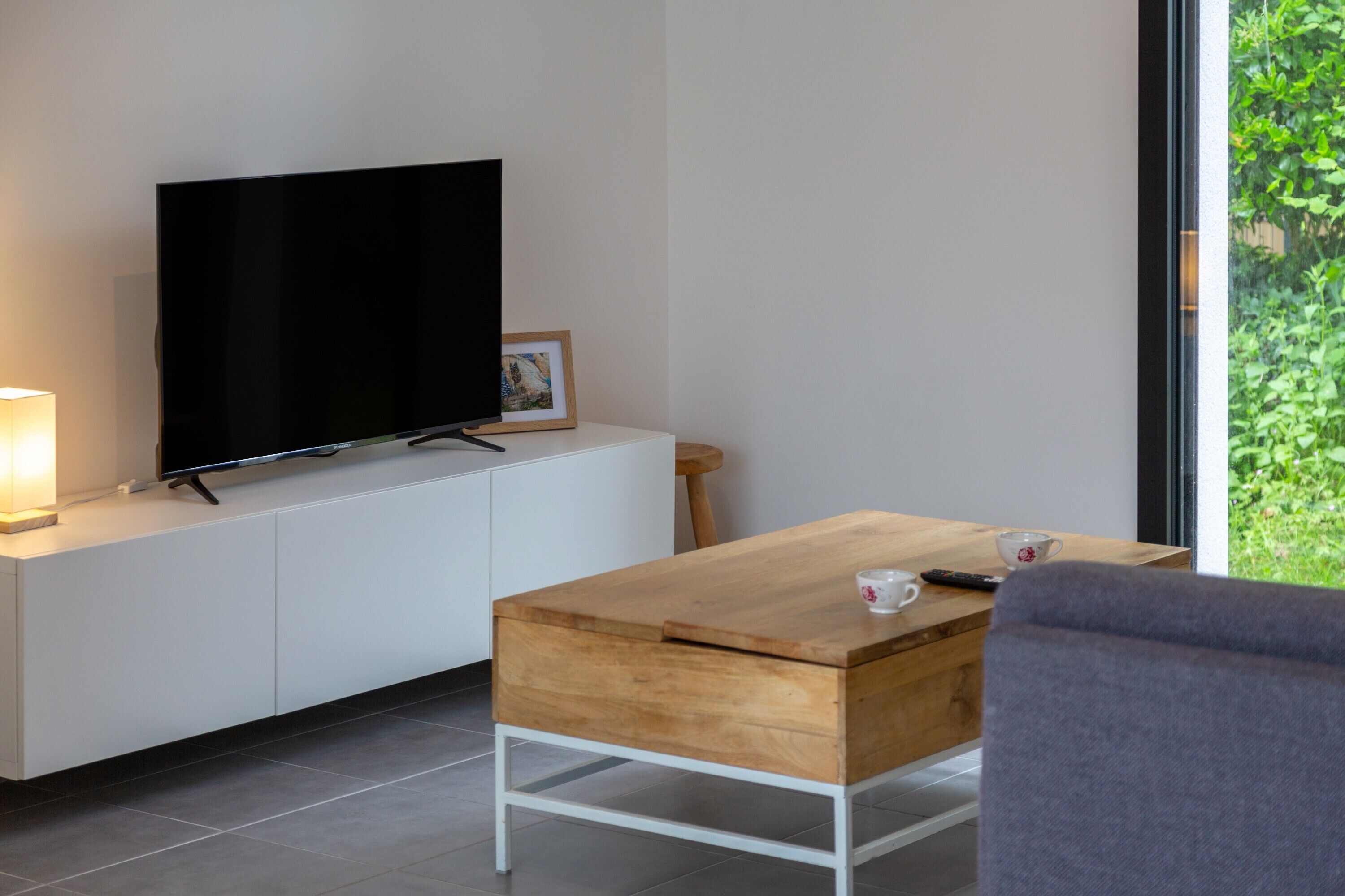 A modern living room with a flat-screen TV on a white cabinet, a wooden coffee table, a grey armchair, and a view of greenery through a large window.