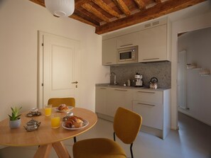 Cocina compacta con microondas, cafetera y lavabo. Hay una mesa de comedor con sillas, una planta en maceta y productos para el desayuno.