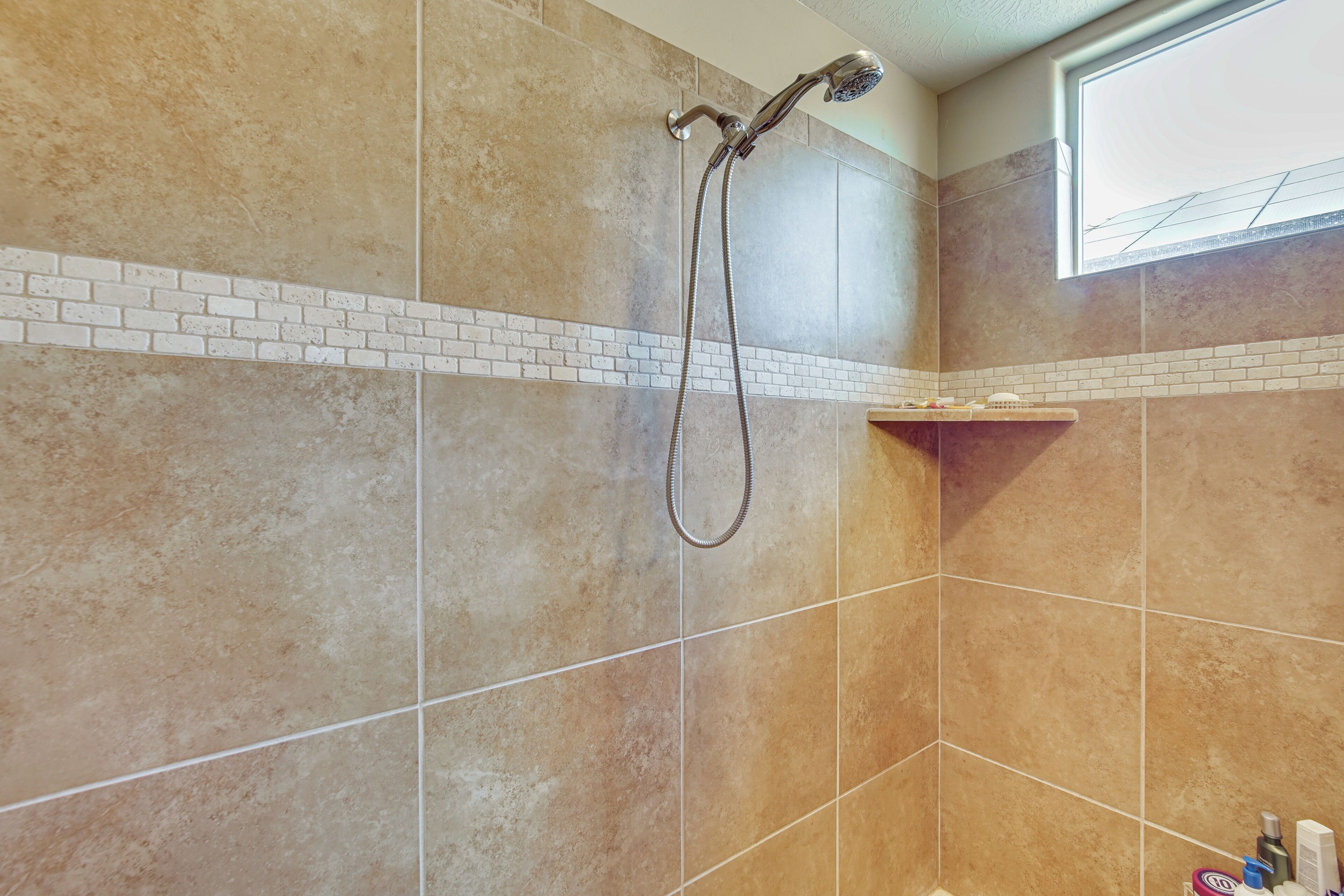 A bathroom with a shower, tiled walls, a window, and a shelf with toiletries.