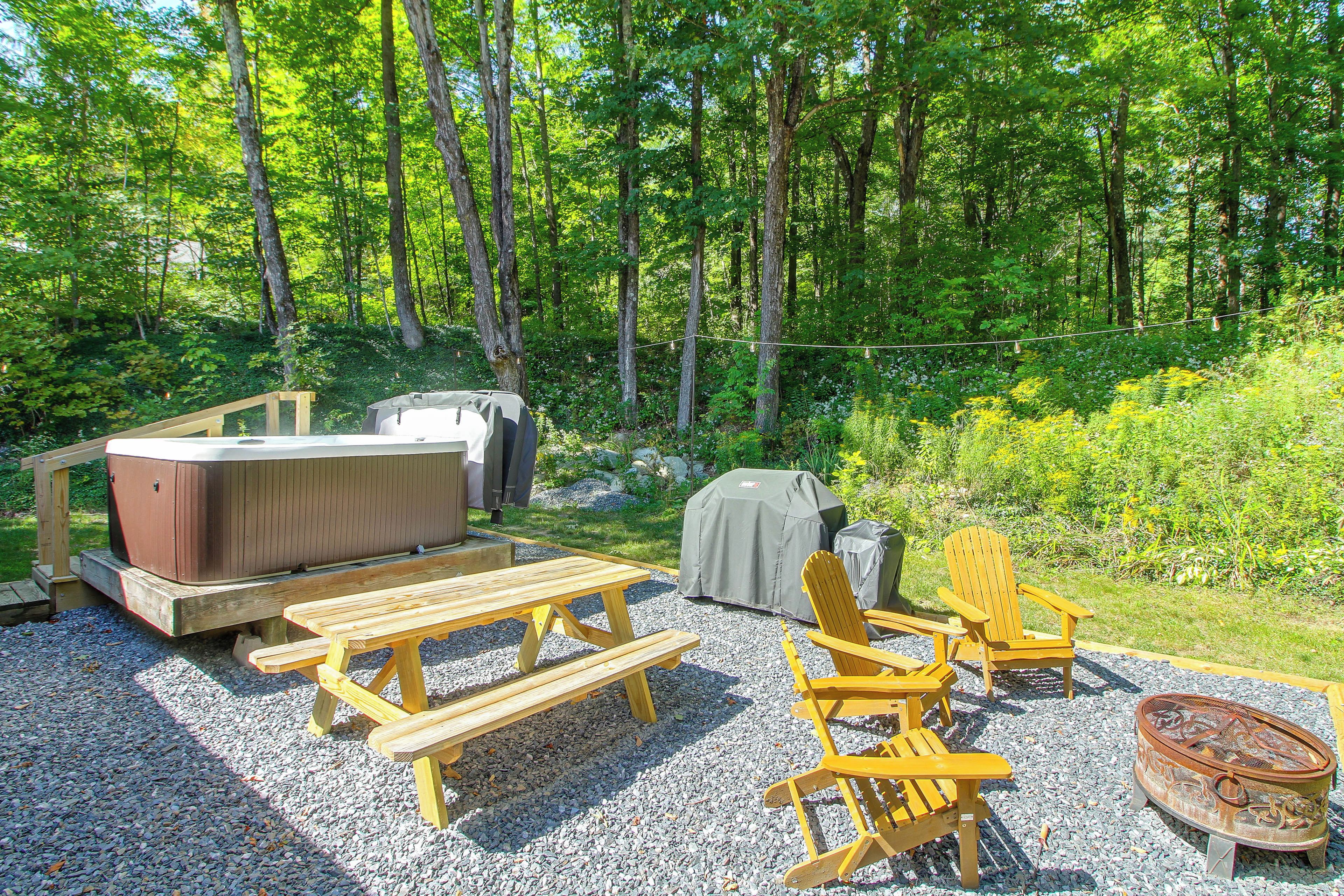 A wooden deck with picnic tables, Adirondack chairs, a hot tub, and a fire pit.