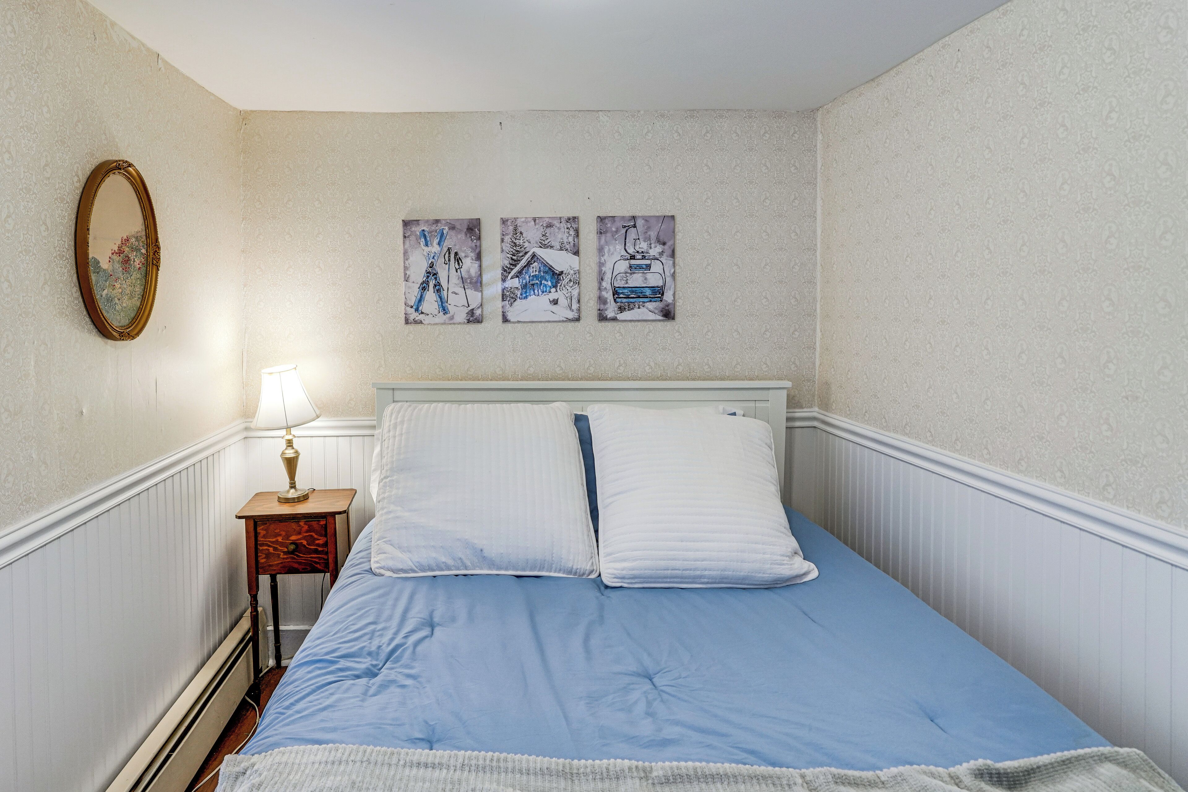 A bedroom with a bed, a nightstand with a lamp, and three wall-mounted pictures depicting winter scenes.