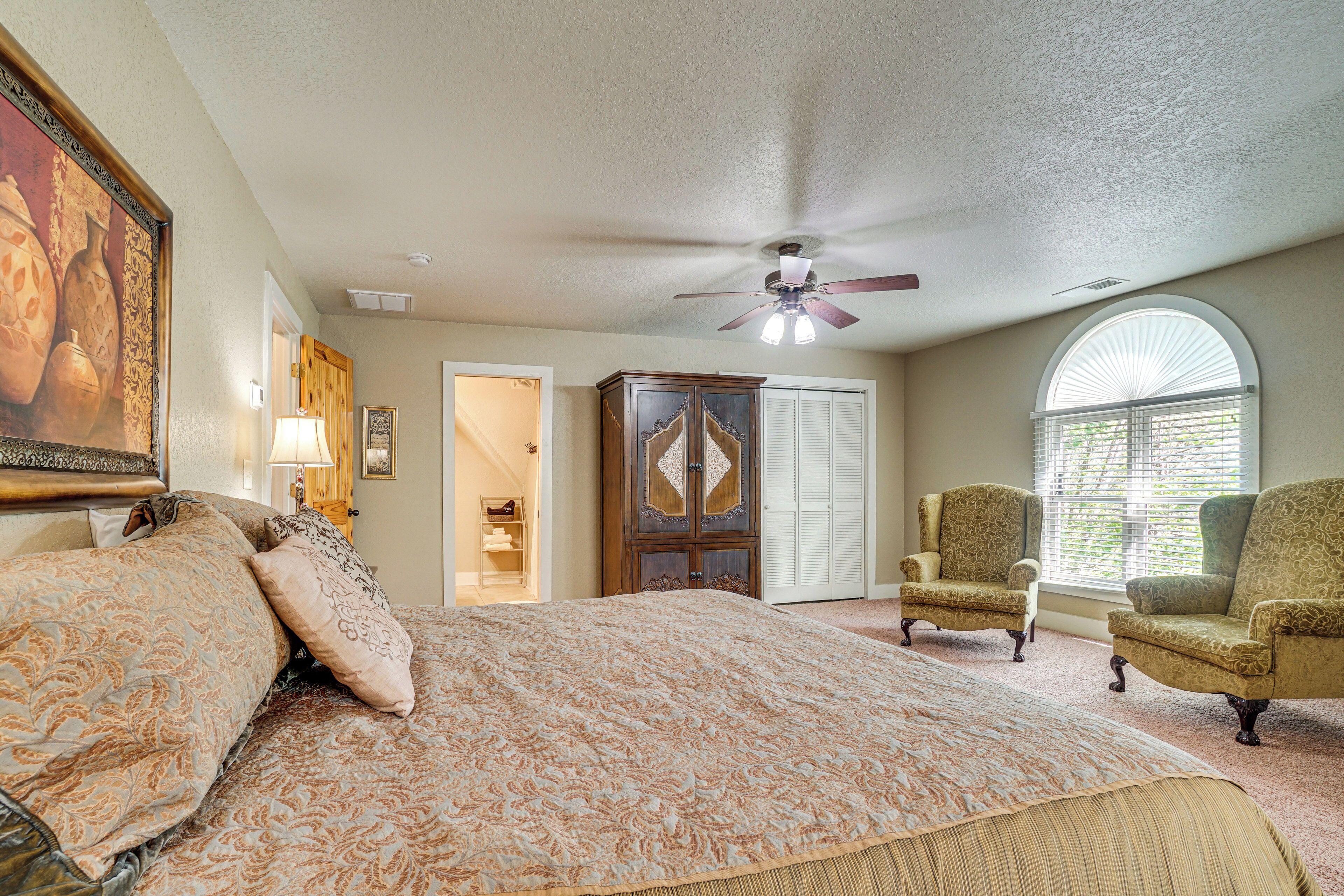 A bedroom with a large bed, a ceiling fan, a wardrobe, two armchairs, and a window with blinds.