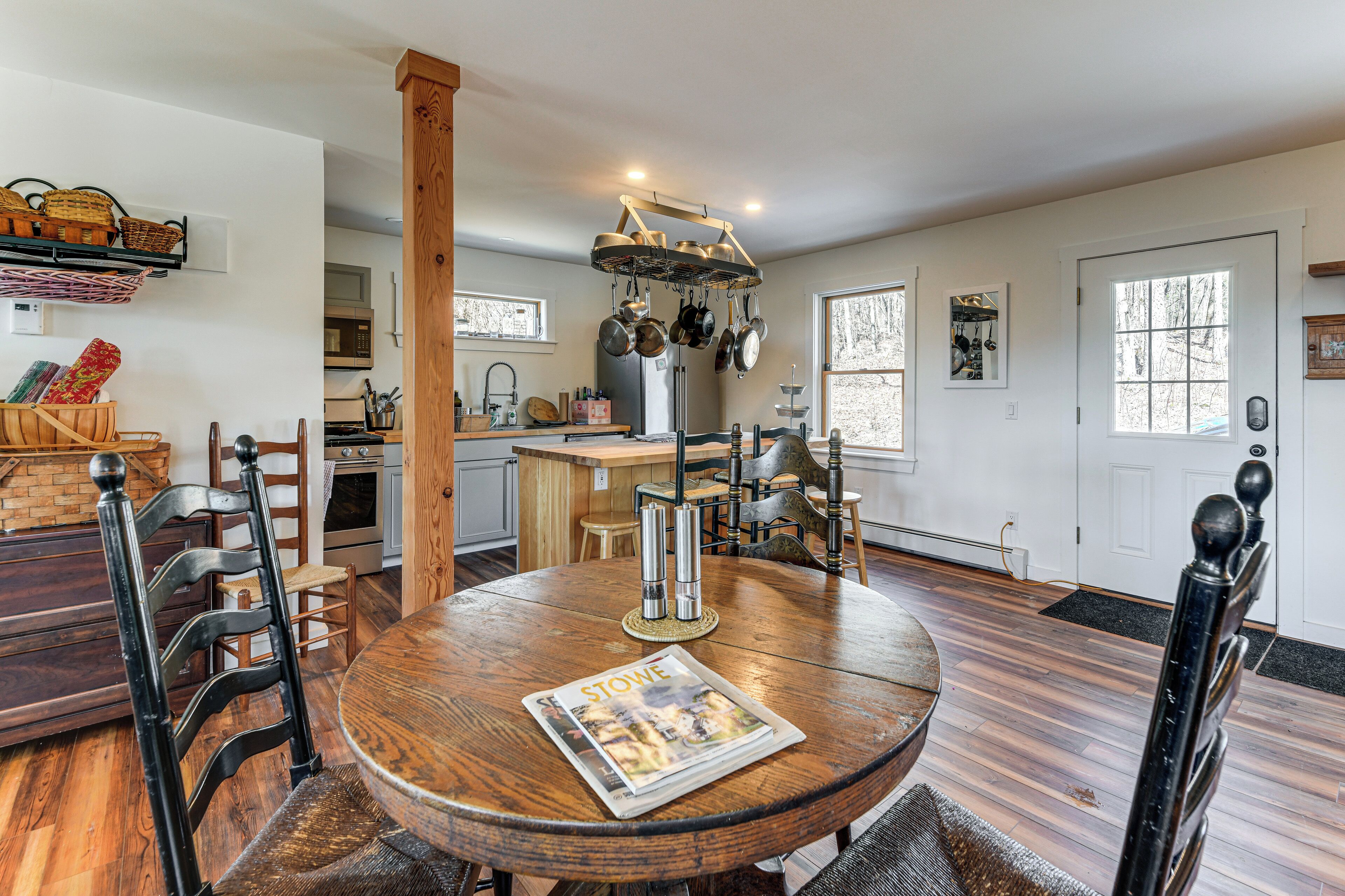 A dining area with a round wooden table, chairs, a hanging pot rack, and a kitchen in the background.