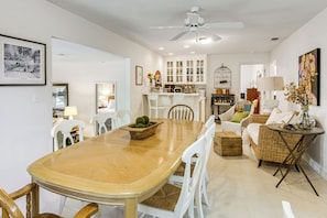 A dining area with a wooden table and white chairs, a white sofa, a wicker chair, a small side table, and a ceiling fan.