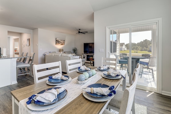 Un coin repas avec une table en bois dressée pour six personnes, des chaises blanches et une vue sur l’extérieur grâce à de grandes portes vitrées.