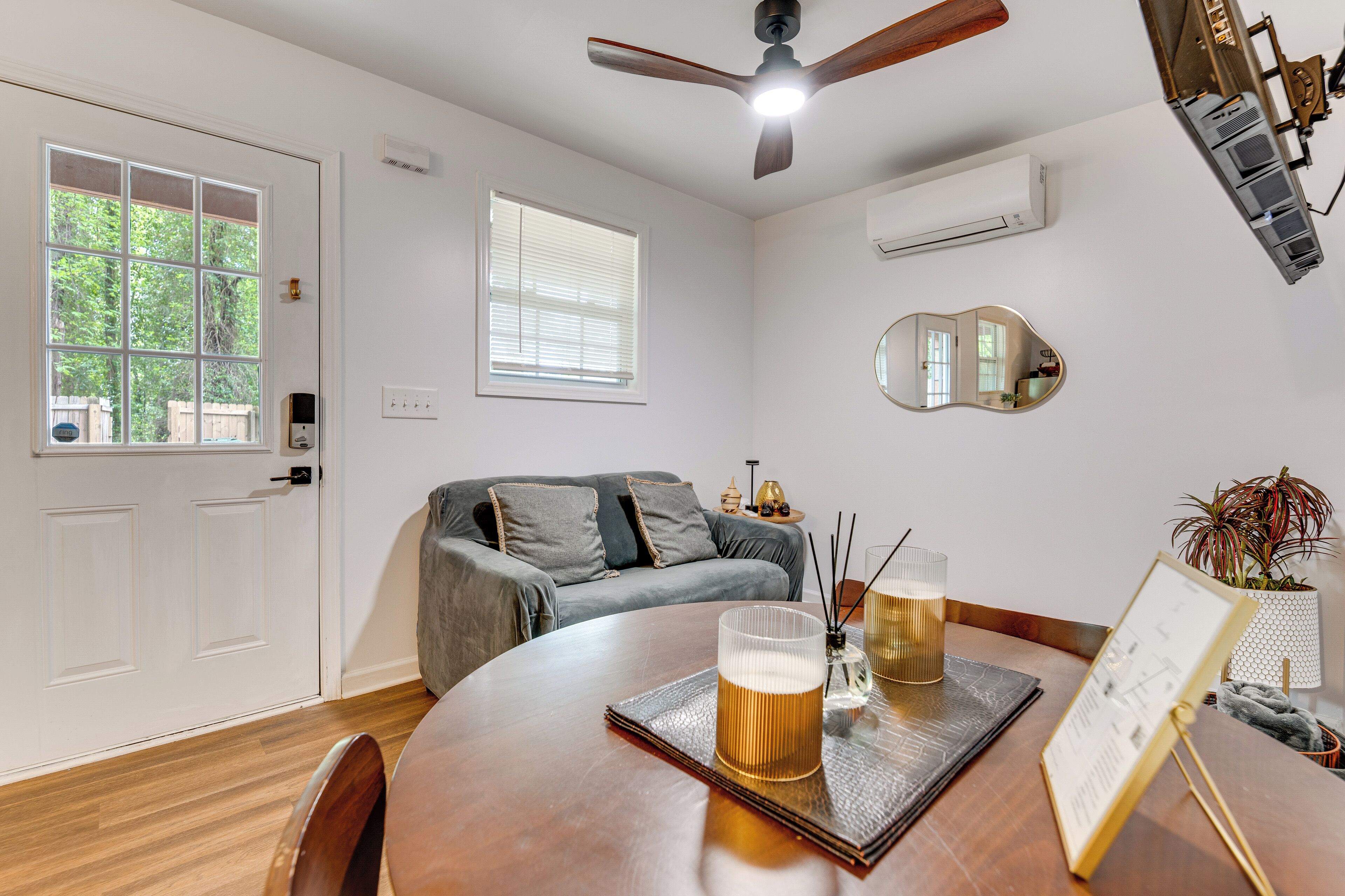 A room with a wooden dining table, a ceiling fan, a window with blinds, a gray sofa, and a wall-mounted air conditioner.