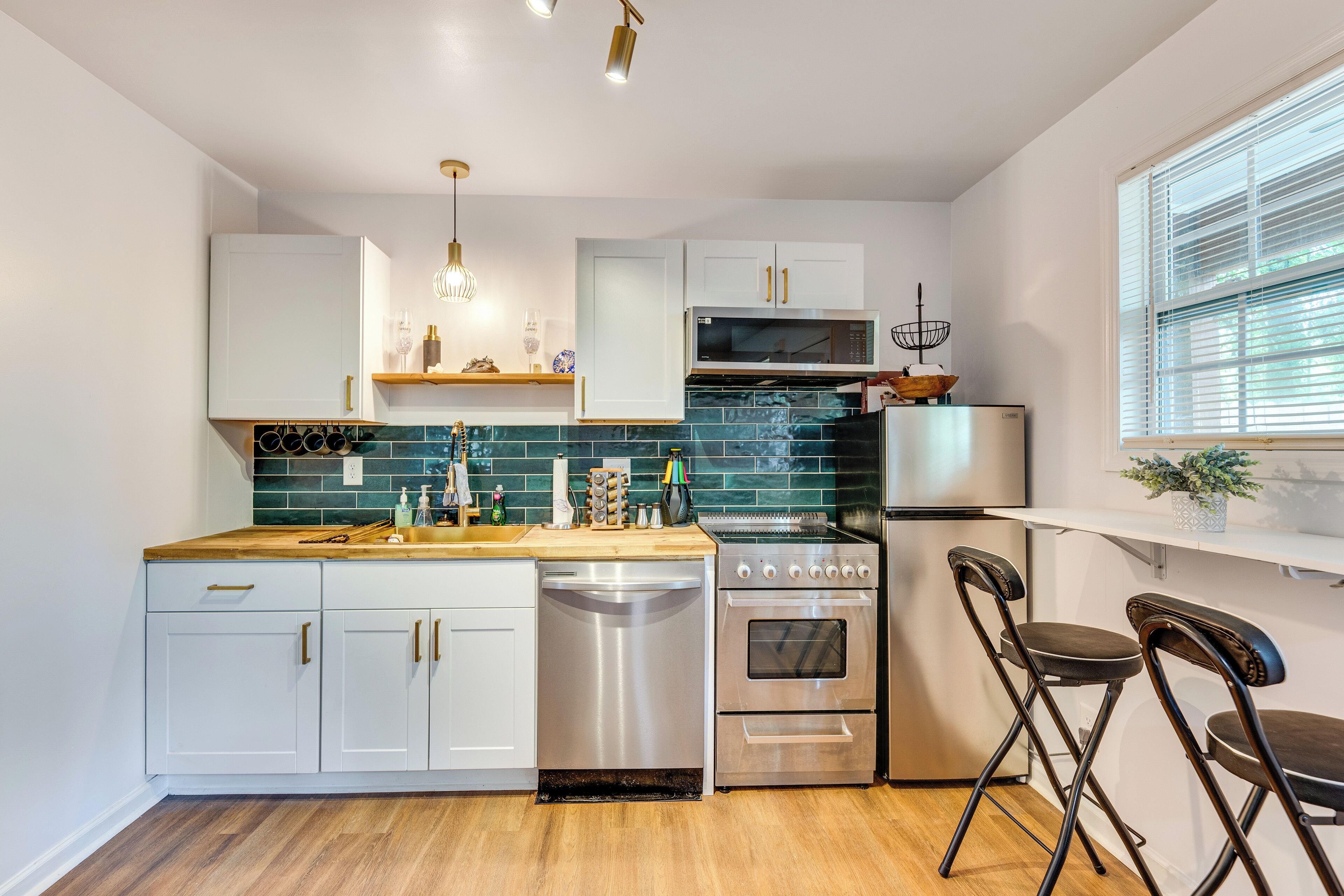 A modern kitchen with white cabinets, a stainless steel dishwasher, a stove, and a microwave. There are two bar stools and a window with blinds.
