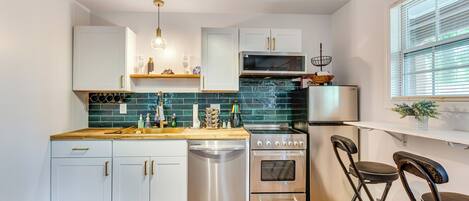 A modern kitchen with white cabinets, a stainless steel dishwasher, a stove, and a microwave. There are two bar stools and a window with blinds.