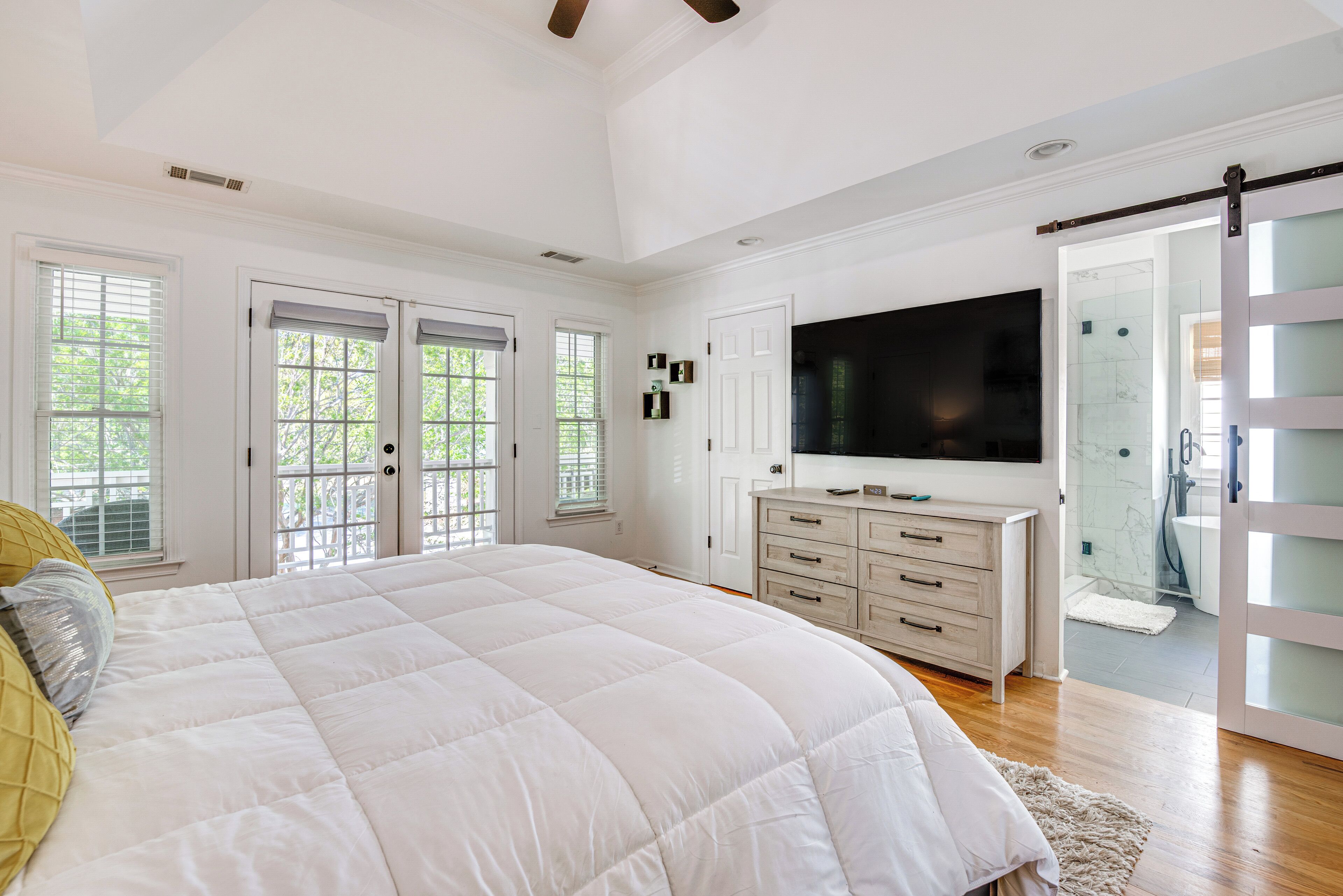 A bedroom with a large bed, a TV, a sliding glass door to the bathroom, and a white color scheme.