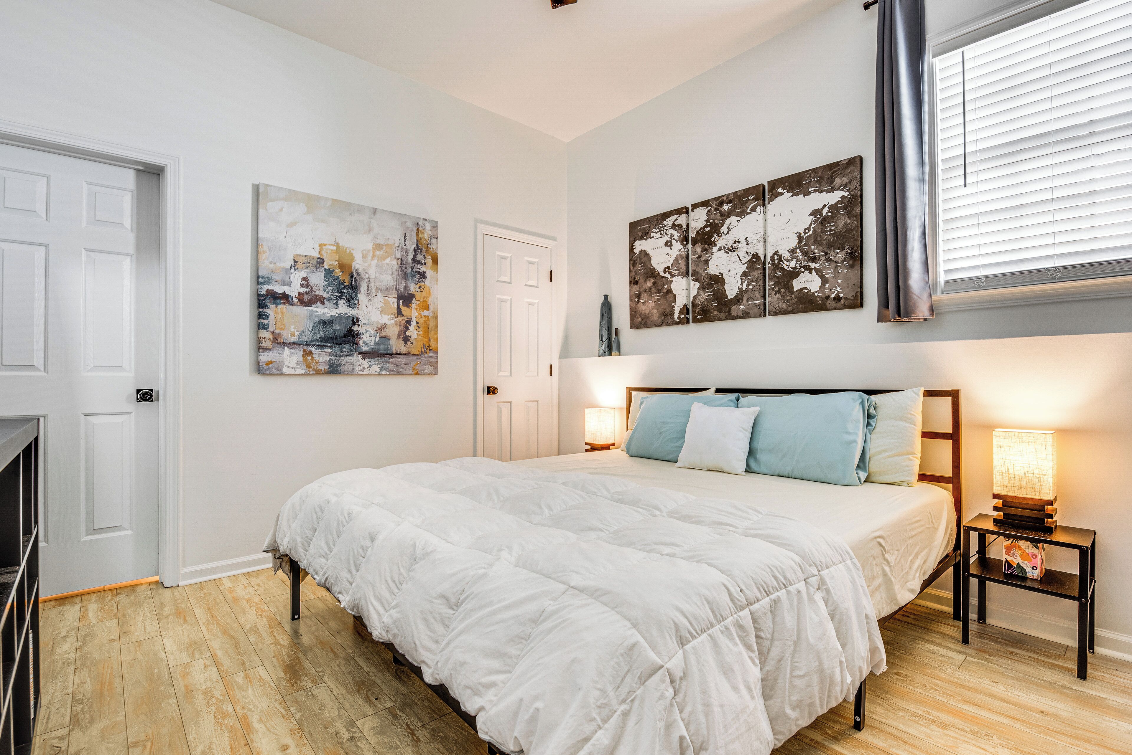 Een slaapkamer met een bed, witte muren, houten vloer, een deur en twee aan de muur hangende schilderijen.