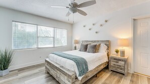 A bedroom with a wooden bed frame, two bedside tables with lamps, a ceiling fan, and a large window with blinds. - Elegant Waterfront Condo w/ Views in Pompano Beach (Pompano Beach)