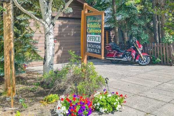 Een houten bord met daarop 'Yellowstone Wildlife Cabins Office' en 'Vakantiewoningen', een rode motorfiets geparkeerd ervoor en een houten hutje op de achtergrond.