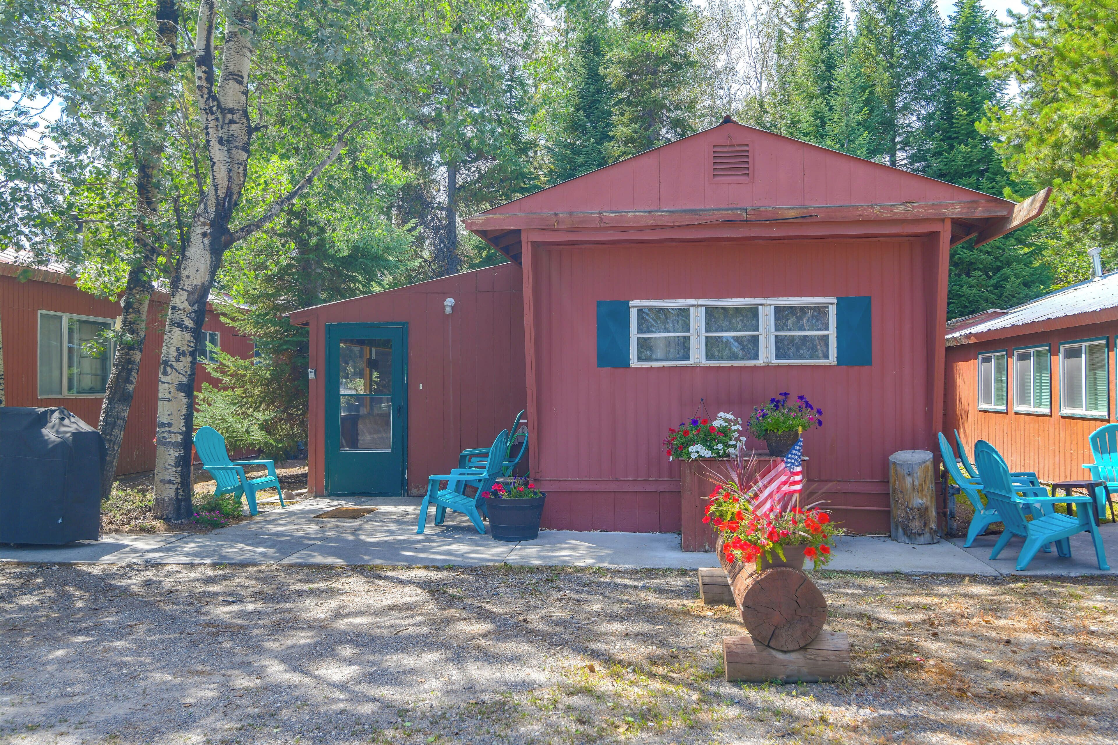 A red building with a door and windows, surrounded by trees. There are blue chairs and a table outside.