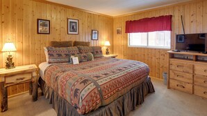 Um quarto com painéis de madeira, uma cama, duas mesas de cabeceira com abajures, um aparador, uma televisão e quadros nas paredes.