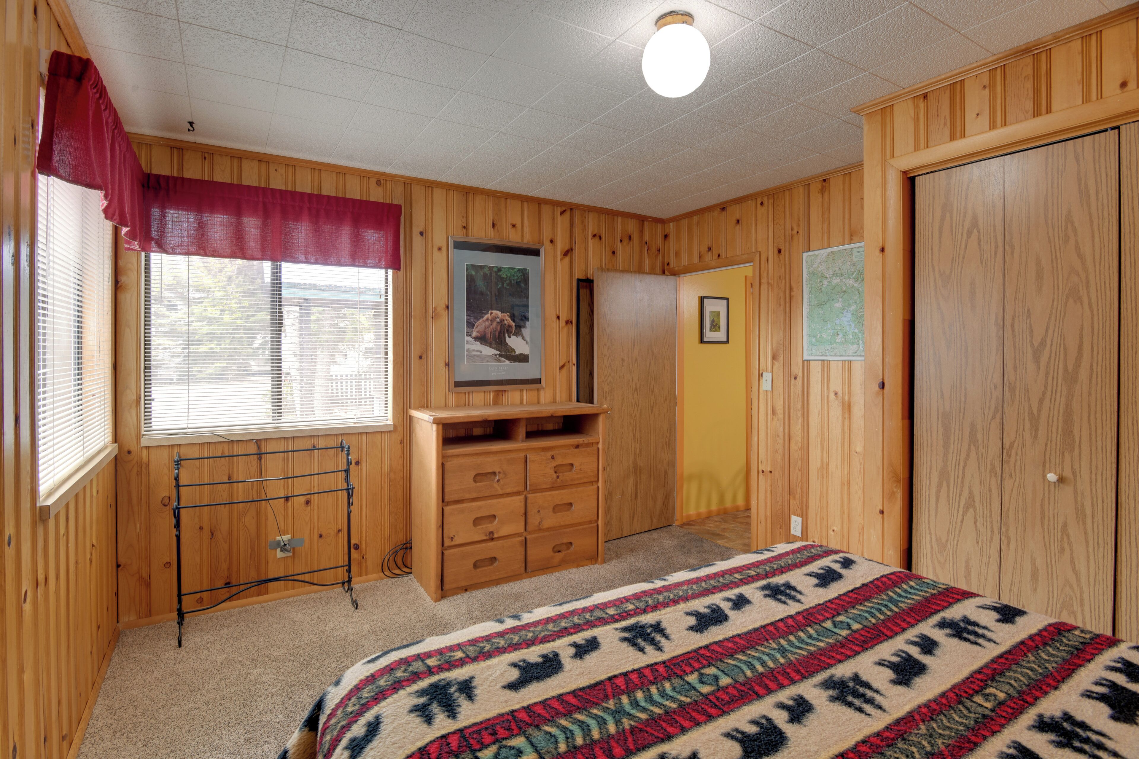 A bedroom with wooden walls, a bed with a patterned blanket, a dresser, a drying rack, and a window with blinds.